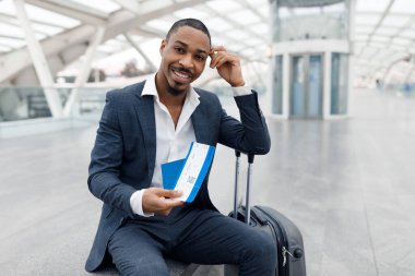Happy Black Businessman Sitting On Bench At Airport, Waiting For His Flight, Handsome Young African American Man Wearing Suit Holding Passport With Tickets And Looking At Camera, Copy Space