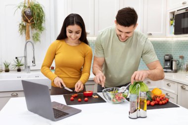 Positive happy young lovers having fun while cooking at home, cheerful hispanic man and woman preparing delicious healthy meal, watching food vlog on Internet, using laptop. Healthy diet