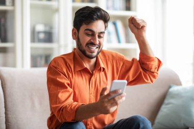 Lottery, giveaway, gambling, online bet. Emotional happy young bearded middle eastern man in smart casual using modern white smartphone, smiling and gesturing, home interior, copy space