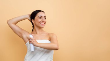 Portrait Of Smiling Young Indian Woman Using Deodorant Spray For Underarms, Beautiful Hindu Female Wrapped In Towel After Bath Using Antiperspirant Aerosol Over Beige Background, Panorama, Copy Space