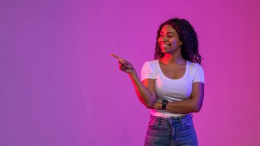 Beautiful Black Woman Standing In Neon Light And Pointing Aside At Copy Space, Smiling African American Female Demonstrating Free Place For Advertisement While Posing Over Purple Studio Background