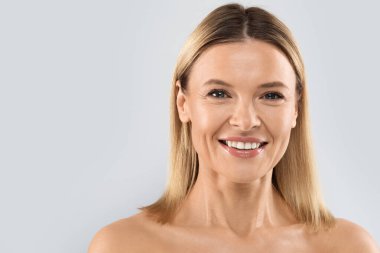 Portrait of happy beautiful half-naked middle aged blonde woman smiling at camera, posing on grey studio background, demonstrating glowing perfect skin, copy space. Cosmetology concept