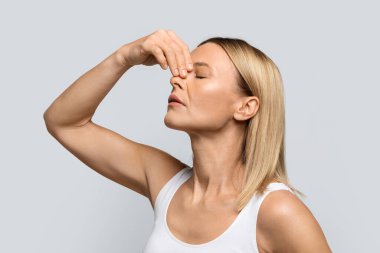 Nosebleeds concept. Middle aged blonde woman with closed eyes touching nose, suffering from bleeding or dry nose, grey studio backgroundm side view, closeup shot clipart