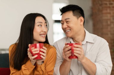 Happy loving japanese spouses sitting on sofa in living room and drinking hot beverage together, looking at each other and smiling, couple enjoying time at home