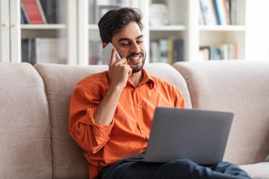 Wealthy cheerful young arab man entrepreneur working from home, sitting on couch, having phone conversation, using laptop, typing on coumputer keyboard, copy space. Remote business concept