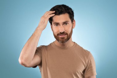Handsome bearded middle aged man in t-shirt touching his hair while posing on blue studio background and smiling at camera, closeup portrait, copy space