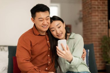 Loving happy korean spouses video calling via cellphone, middle aged man and his young wife relaxing on sofa at home, browsing new app or shopping online