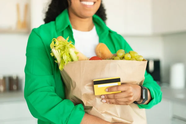 Grocery Shopping. Unrecognizable African American Lady Holding Credit Card And Paper Bag With Food Products Standing In Modern Kitchen At Home. Cropped, Selective Focus