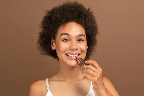 Glad young black curly female with perfect skin applying lipstick on lips for moisturizing or nude makeup isolated on brown background, studio. Ad and offer, cosmetics treatment and beauty lesson