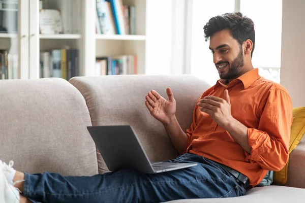 Cool middle eastern young handsome guy in smart casual outfit chilling on couch in living room, using laptop, arab man have video call with lover or family while resting at home, copy space