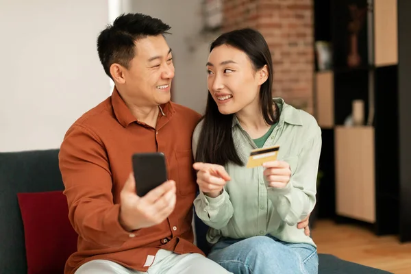 Banking application and e-commerce concept. Excited asian spouses using smartphone and credit card, making online payment or shopping, sitting on sofa at home