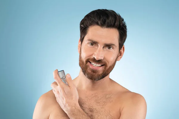 Happy bearded man spraying on cologne and smiling at camera, doing morning routine over blue studio background. Beauty skin care, moisturizers products and perfume