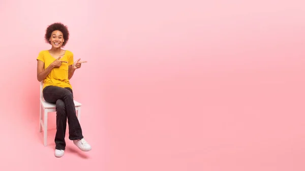 Smiling pretty young black curly female sitting on chair, pointing fingers at empty space isolated on pink background, panorama, studio. Great news, recommendation, advice for sale, ad and offer