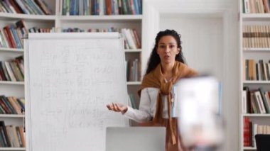 Online math lesson. Young woman tutor recording video lesson on smartphone for students, explaining mathematics with book, talking to smartphone, tracking shot