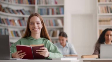 Happy studentship concept. Positive carefree young woman student reading book and smiling to camera, studying in campus library with groupmates, slow motion, empty space