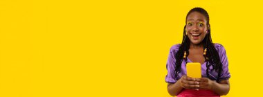 Mobile Offer. Excited Black Lady Holding Cellphone Looking At Camera Standing Near Copy Space Over Yellow Studio Background. New Application Advertisement. Panorama