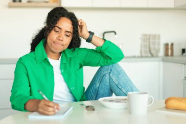 Black Woman Learning And Having Breakfast Taking Notes Sitting In Modern Kitchen At Home. Pensive Lady Writing Planning Healthy Diet Menu. Dieting And Nutrition Concept