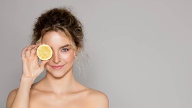 Beautiful woman holding lemon half and looking at camera, lady using vitamin c fruit acids for beauty care, posing over grey background, panorama with copy space