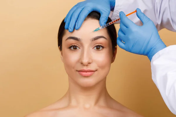 Injection Cosmetology. Beautician Doctor Making Botox Shot To Young Indian Woman Forehead, Attractive Hindu Lady Getting Anti-Wrinke Treatment While Standing Over Beige Studio Background, Copy Space