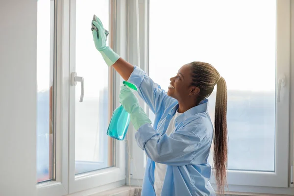 Professional housekeeping, cleaning service. Happy cheerful attractive young black woman housekeeper cleaning windows at apartment, using spray and cloth, wearing rubber gloves, side view, copy space