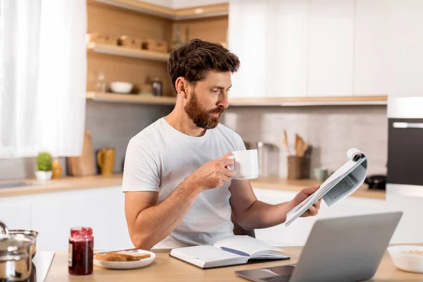 身穿白色T恤的活泼迷人的成熟的高加索人与图表一起工作 在现代厨房的内部与笔记本电脑一起喝咖啡 远程办公 早上好 新的正常生活 — 图库照片