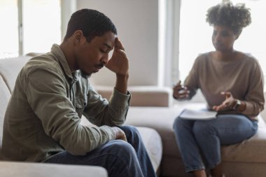 Upset millennial black guy sitting on couch with closed eyes, touching head, suffering from depression, mental disorder, visiting psychotherapist, african american lady counselor talking and gesturing
