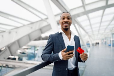 Handsome Young Black Businessman Using Smartphone And Credit Card While Standing In Airport, Smiling African American Male Wearing Suit Making Mobile Payments While Waiting Flight, Copy Space