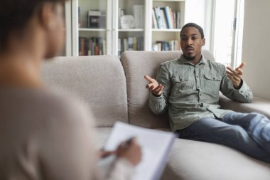 Emotional african american millennial guy in casual reclining on couch at therapist office, sharing feelings and gesturing, young black man having therapy session with woman psychologist, free space