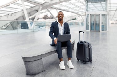 Remote Business. Smiling Black Businessman Working On Laptop While Waiting At Airport, Young African American Male Entrepreneur Sitting On Bench In Terminal And Using Computer For Online Work