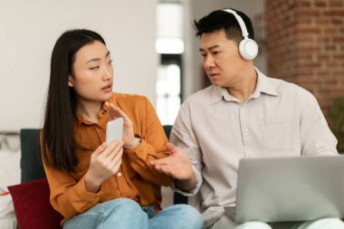 Marital problem. Asian middle aged husband looking at wifes smartphone while using laptop and wearing headphones, sitting together on sofa