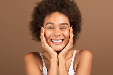 Glad smiling young black curly female with perfect skin presses her hands to face, enjoys fresh isolated on brown background, studio. Cosmetics for nude makeup, beauty care and cleansing, treatment