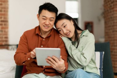 Happy korean spouses using digital pad, surfing internet or shopping online, sitting on sofa at home interior, free space. Middle aged man and his young wife using modern gadget