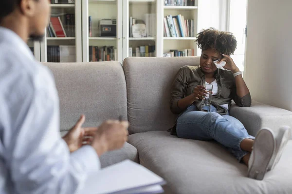 Crying broken young african american woman in casual outfit reclining on sofa at psychologist office, having therapy session. Black man psychotherapist helping depressed lady with PTSD, copy space