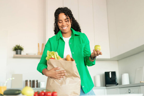Smiling Black Lady Unpacking Paper Grocery Bag After Shopping Holding Fresh Apple Standing Near Table In Modern Kitchen At Home. Household And Dinner Preparation Concept