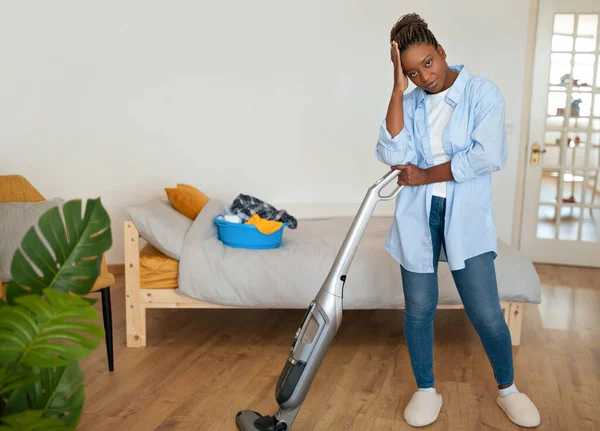 Exhausted unhappy young black woman in homewear housewife cleaning house alone, using modern vacuum cleaner, feeling tired, depressed, copy space for cleaning housekeeping service ad