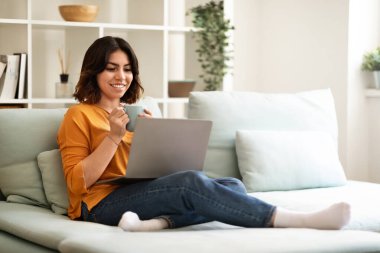 Domestic Pastime. Happy Young Arab Female Relaxing With Laptop And Coffee At Home, Smiling Middle Eastern Woman Sitting On Couch, Watching Movies On Computer And Enjoying Hot Drink, Copy Space