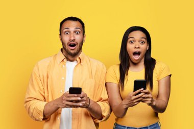 Glad shocked young black couple with open mouth chatting on phones isolated on yellow background, studio. Surprise, reaction to news, relationship, love, gadget for social network, blog, ad and offer