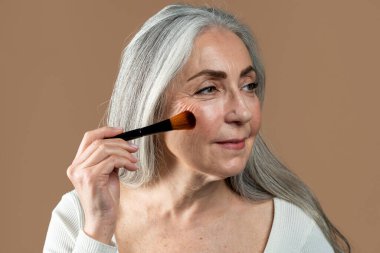 Glad old european lady with gray hair applies powder, blush with brush on her face, isolated on brown background, studio, close up. Anti-aging cosmetics and nude makeup, beauty care, ad and offer