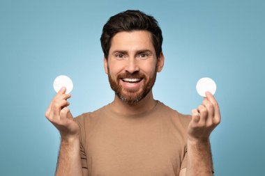 Portrait of excited bearded man with perfect skin holding in hands cotton pads and smiling at camera, posing over blue background. Skin care healthcare cosmetic procedures concept