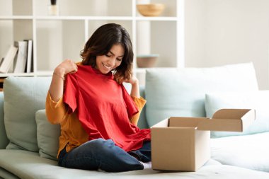 Smiling Young Middle Eastern Female Unpaking Box With New Clothes At Home, Happy Arab Woman Taking Out Red T-Shirt, Checking Item After Parcel Delivery, Satisfied With Online Shopping, Copy Space