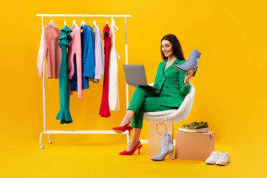 Young lady shop assistant holding elegant shoe and using laptop, sitting near clothing rail and receiving online orders, selling outfits over internet on yellow studio background