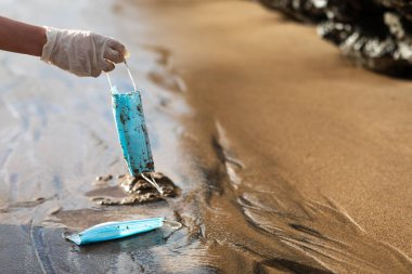 Female hand picking up used disposable medical face masks on the beach while cleaning the coastal zone, free space. Plastic pollution, medical waste, garbage, trash on sea water