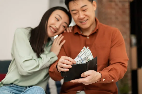 Positive rich middle aged asian man putting banknotes into wallet and smiling, sitting with his young wife on sofa at home, selective focus. Finance and money saving concept