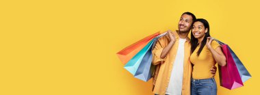 Satisfied young black couple hold many packages with purchases, look at empty space for ad and offer, isolated on yellow background, studio, panorama. Shopping together, sale and buyers enjoy buy