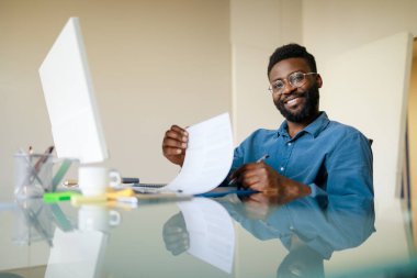 Happy black businessman working with documents, taking notes to financial report or signing contract, male entrepreneur holding pen in hand while sitting at desk in modern office