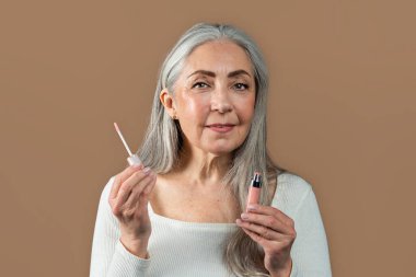 Smiling old european lady with gray hair shows lip gloss and brush, recommend ad and offer, isolated on brown background, studio. Daily procedure, anti-aging cosmetics, nude make-up and beauty care