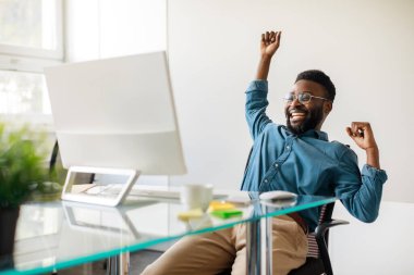 Great news concept. Excited black businessman celebrating online win sitting at desk, looking at computer monitor, copy space. Happy african american got promotion, raising hands up