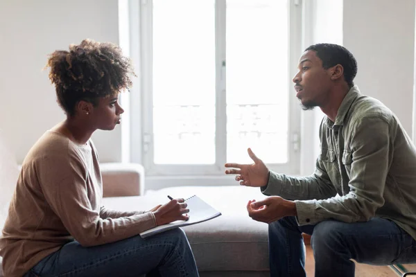 Handsome young black man in casual sitting on couch at counselor office, sharing feelings, thoughts with therapist african american woman, having therapy session, copy space. Mental dosirders concept