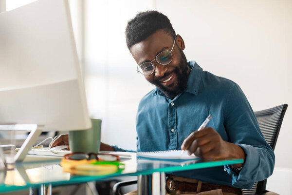 African american male entrepreneur working on modern computer and taking notes, sitting at workpace in office interior. Entrepreneurship career, lifestyle and occupation concept