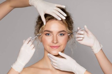 Attractive young woman getting injection for eyes area by two cosmetologists, beautician hands in gloves giving anti aging serum shot on female face against grey background